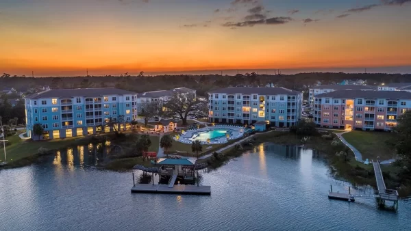 Aerial shot of the apartment community taken from above the water with the illuminated pool and apartment buildings at sunset