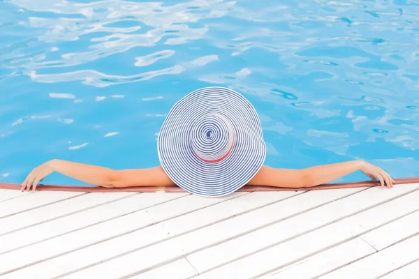 Woman with her back towards the camera resting at the edge of the pool wearing an oversized beach hat